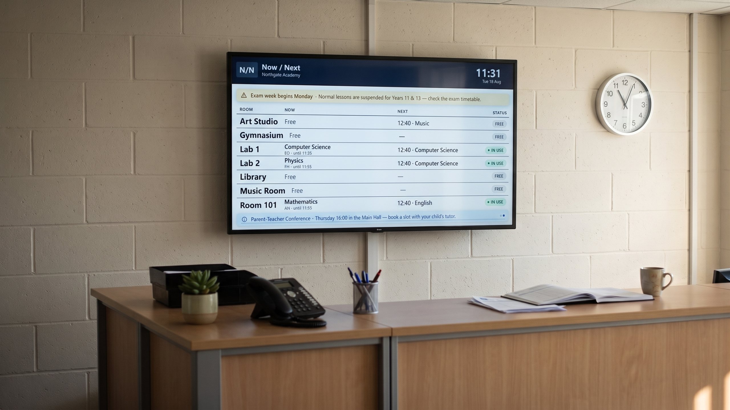 The room board on a screen above a school reception desk, a phone, a plant and a wall clock reading the same time.